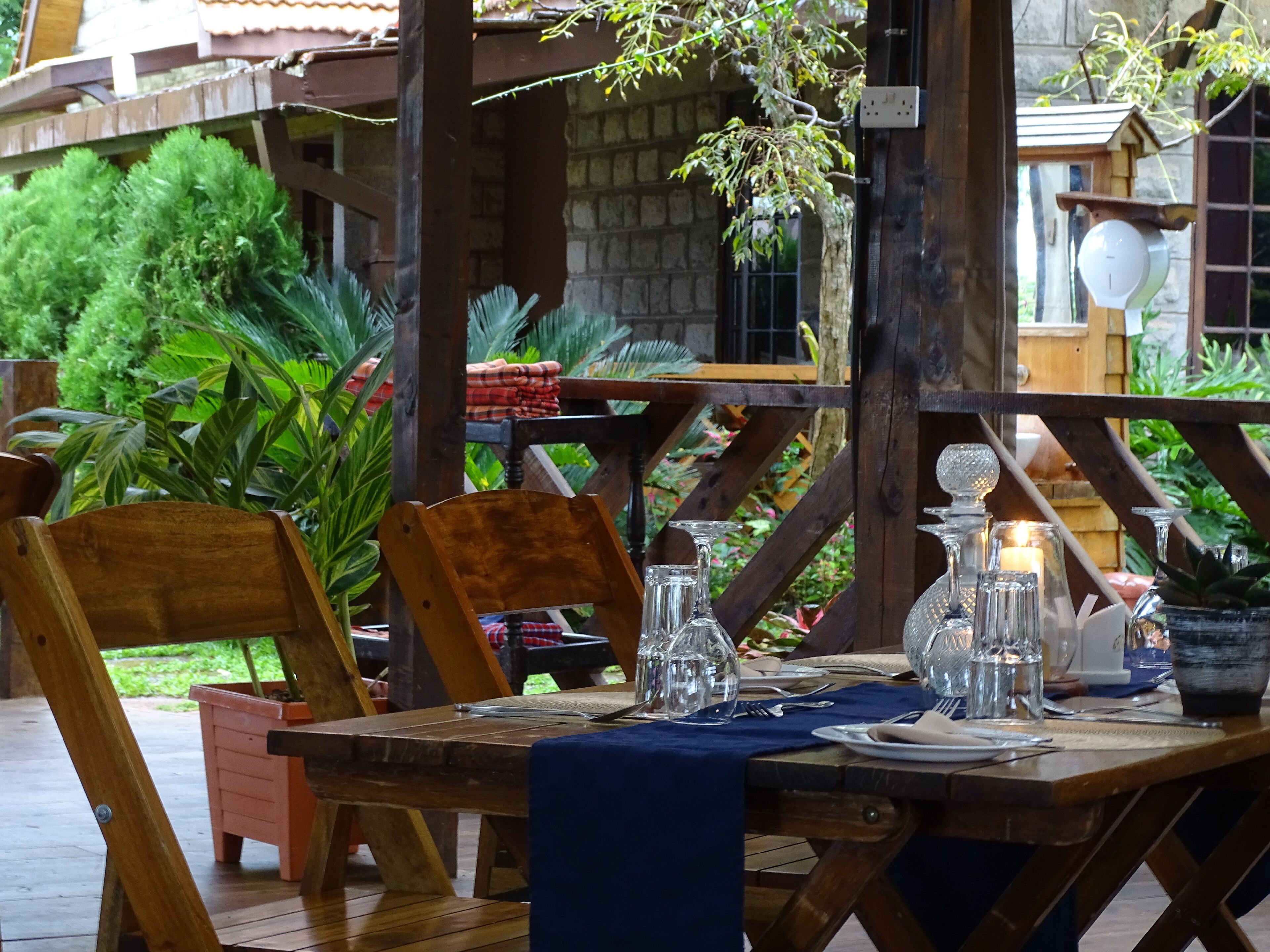 An outdoor wooden table set with glassware and a blue tablecloth, surrounded by wooden chairs and potted plants.