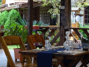 Una mesa de madera al aire libre con cristalería y un mantel azul, rodeada de sillas de madera y plantas en macetas.