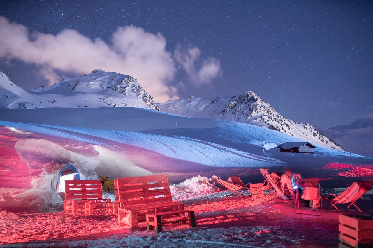 A snowy mountain landscape with a clear sky, a small cabin, and red seating areas.