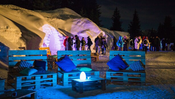 Een besneeuwde buitenruimte met houten banken en tafels, waarvan sommige kussens hebben.