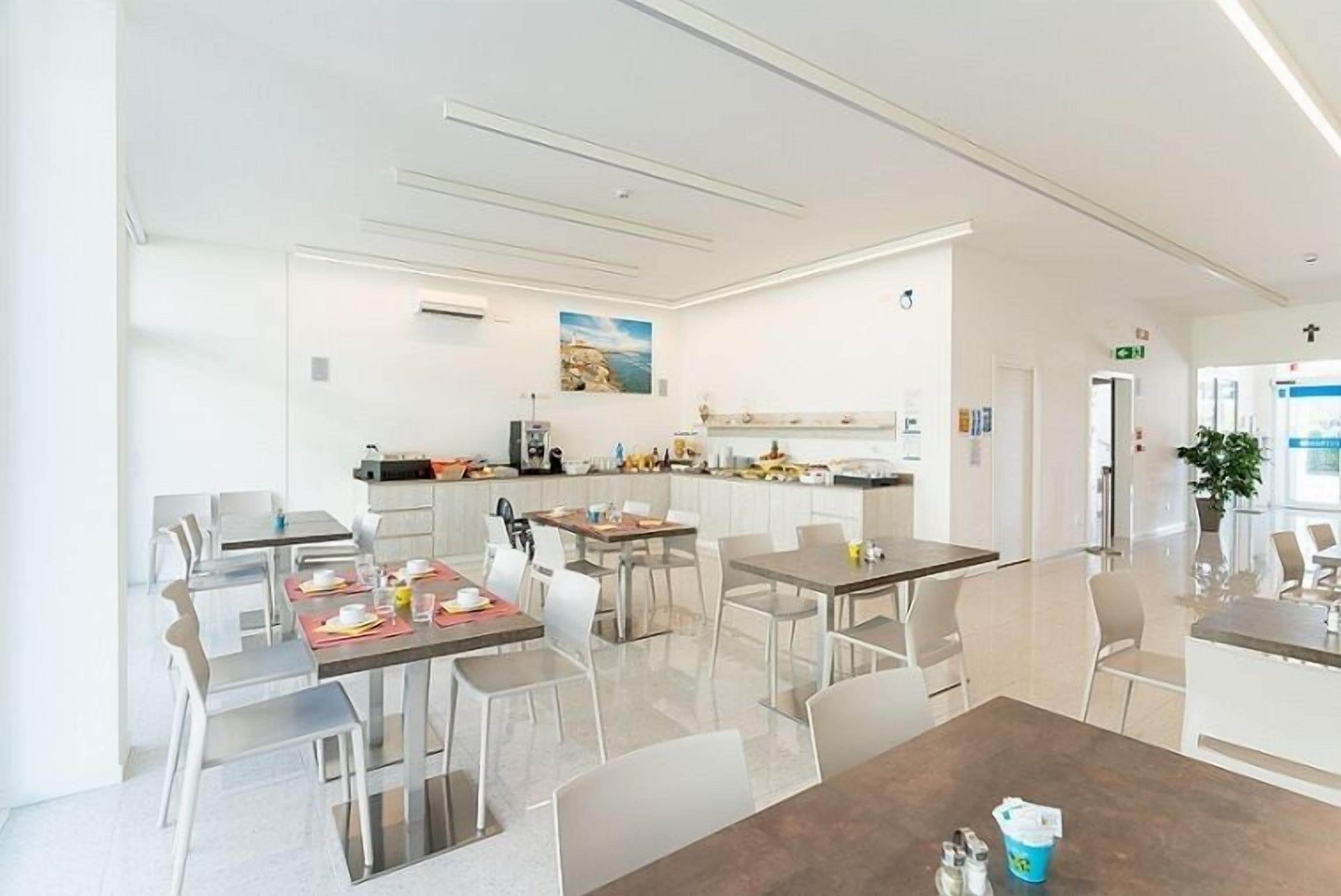 Un comedor moderno con varias mesas y sillas, una cocina al fondo, y un interior limpio y blanco.