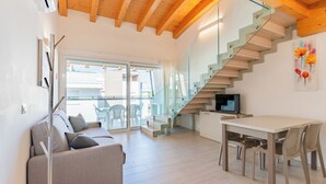 A modern living room with a sofa, dining table, chairs, a TV, and a staircase. - Residenza Turistica Alberghiera Blue Marine (San Michele al Tagliamento)