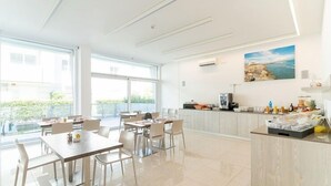 A bright, spacious dining area with large windows, a kitchen counter with various items, and a painting of a coastal scene. - Residenza Turistica Alberghiera Blue Marine (San Michele al Tagliamento)