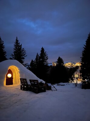 Un igloo de neige doté d’une porte et de bancs à l’extérieur, entouré d’arbres et avec une montagne en arrière-plan.