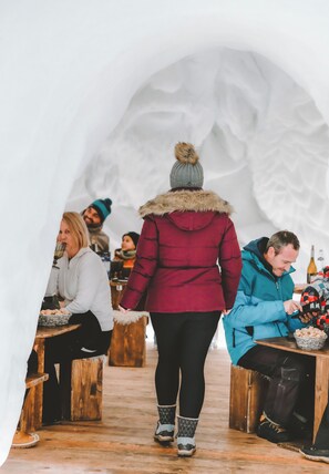 Un espace intérieur avec des tables et des bancs en bois, une personne portant une veste rouge avec une capuche bordée de fourrure, et une autre personne portant une veste bleue.