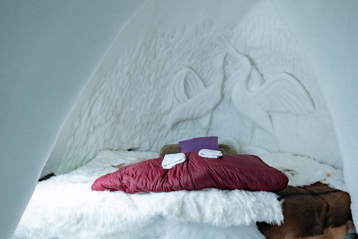 Een gezellig, iglo-vormig bed bedekt met een sneeuwwitte stof, met een zacht rood kussen en een paars kussen, tegen een witte muur met een structuur.