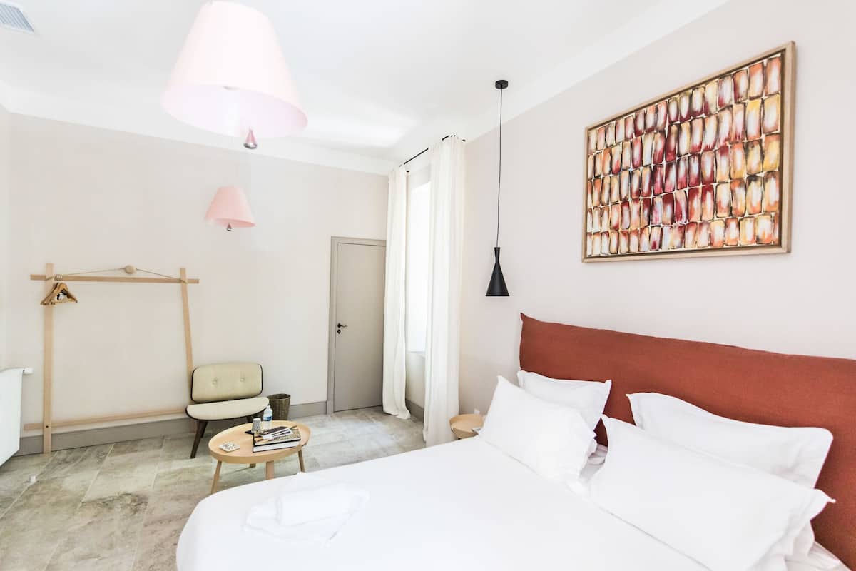 Quarto de hotel com cama, uma cadeira, uma mesinha, uma porta e uma obra de arte na parede.