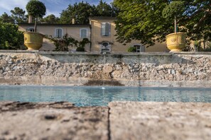 A stone wall with a clear pool, two large yellow pots, and a building with windows and shutters in the background.