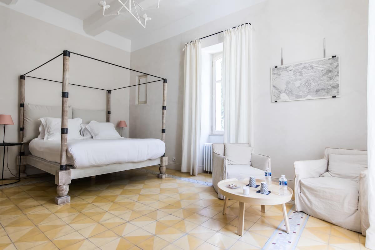 Quarto com cama de dossel, uma mesa redonda com xícaras e garrafas, e uma cadeira.