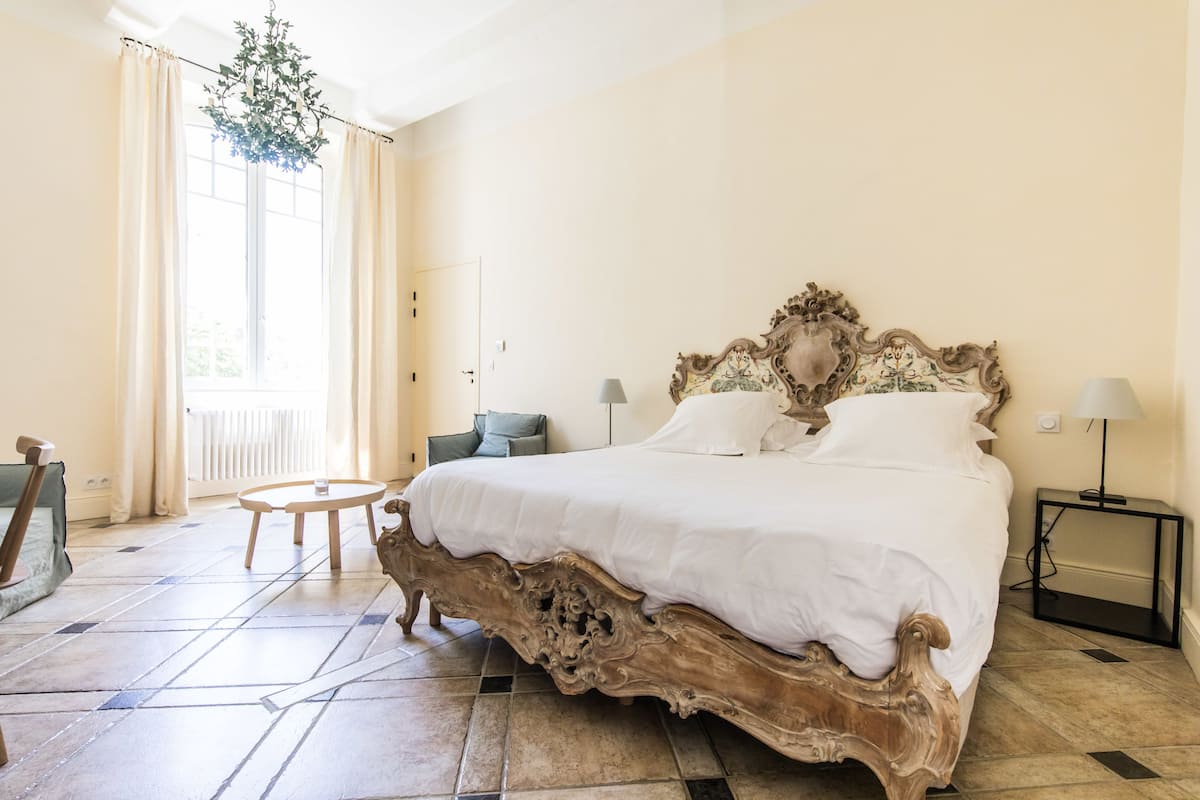 Um quarto espaçoso com uma cama grande e ricamente decorada, uma mesa de madeira redonda e vista para o exterior pela janela.