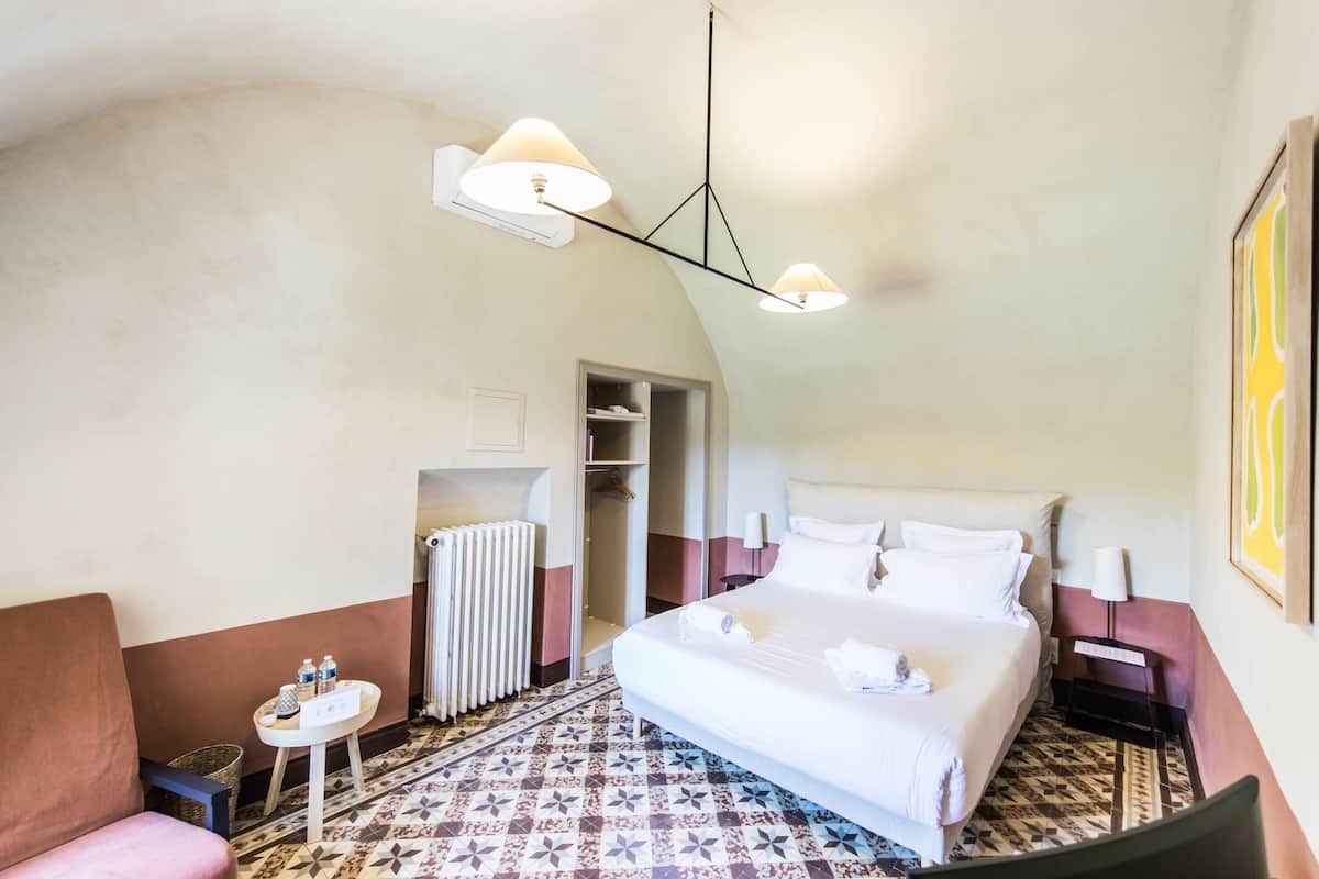 Quarto de hotel com uma cama grande, uma mesinha com abajur, radiador e armário.