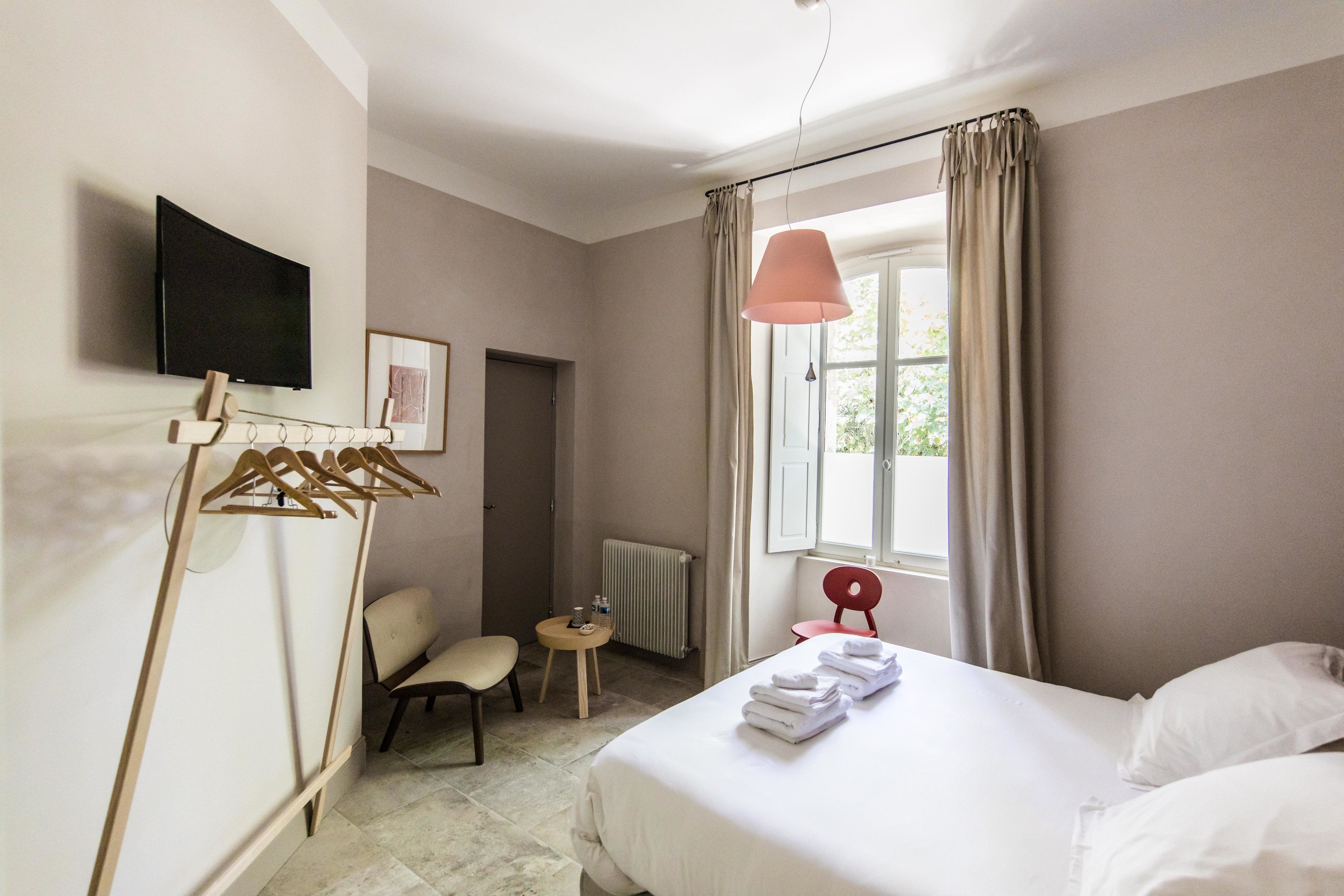 Ein Hotelzimmer mit einem Bett, einem Sessel, einem kleinen Tisch mit einer Flasche, einem an der Wand befestigten Fernseher und einem Fenster mit Vorhängen.