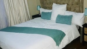 A neatly made bed with a tufted headboard, white and teal bedding, and a teal throw blanket. There are two bedside tables with lamps. - Royaltics Place (Randburg)