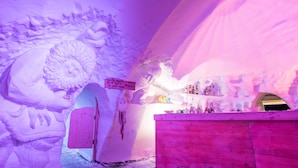 Interior de un hotel de hielo con paredes de hielo talladas, una zona de bar y un cartel que indica un tour guiado.