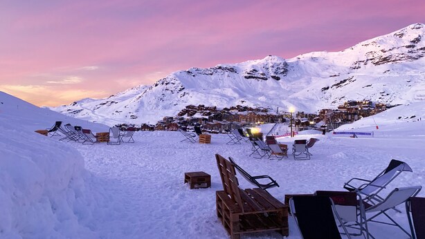 Un paisaje montañoso nevado con un pueblo al fondo, zonas de estar al aire libre con sillas y mesas, y un cielo despejado.