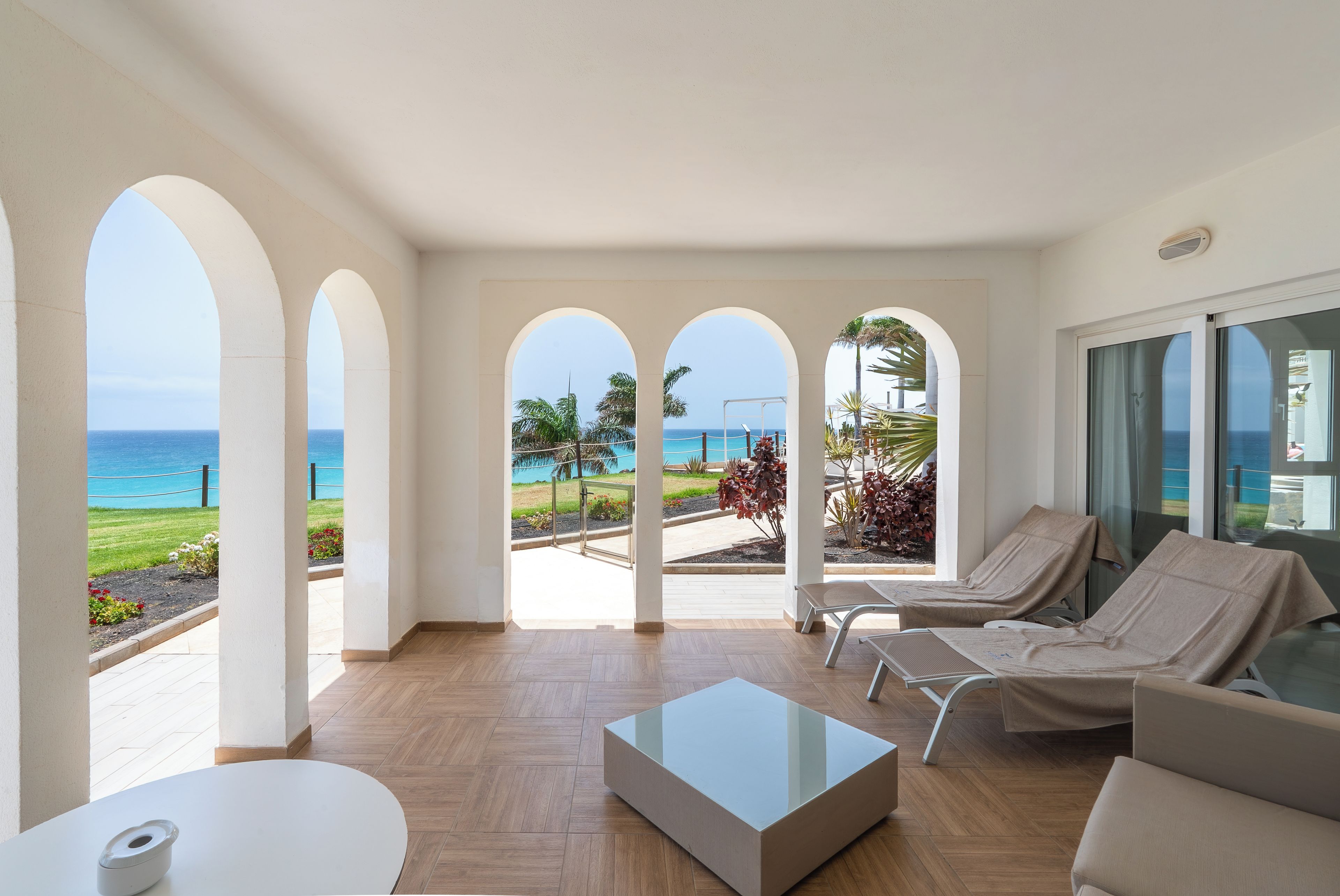 A hotel room with a view of the sea, a balcony with lounge chairs, and a glass coffee table.