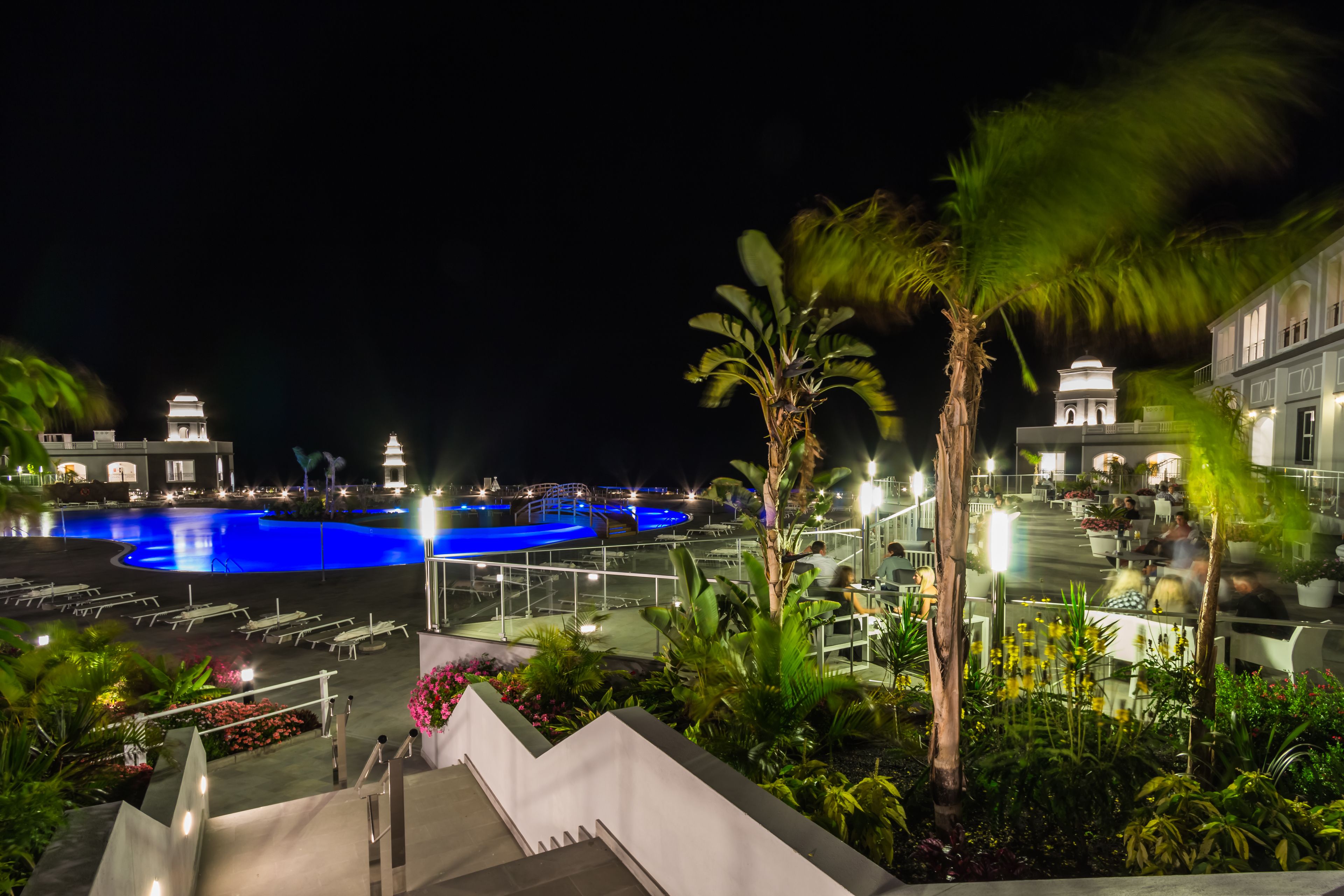 Een nachtzicht van een hotel met een zwembad, terras en palmbomen.