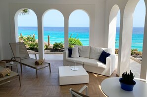 Une pièce baignée de lumière, avec un canapé blanc, un fauteuil et une table, donnant sur une mer turquoise à travers de grandes fenêtres en arc de cercle.