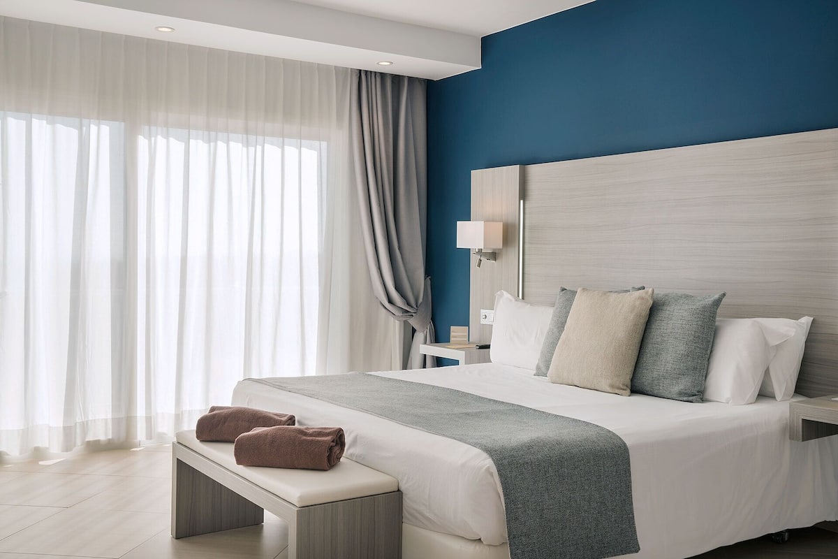 A modern hotel room with a large bed, a bedside table, a nightstand, a lamp, and a bench with towels.