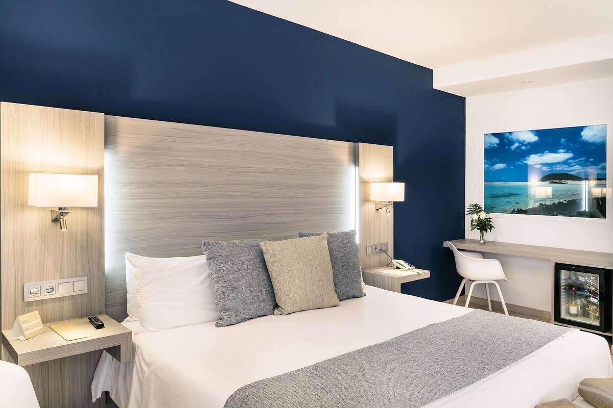 A modern hotel room with a large bed, two bedside lamps, a nightstand, a desk with a chair, and a framed picture of a beach scene.