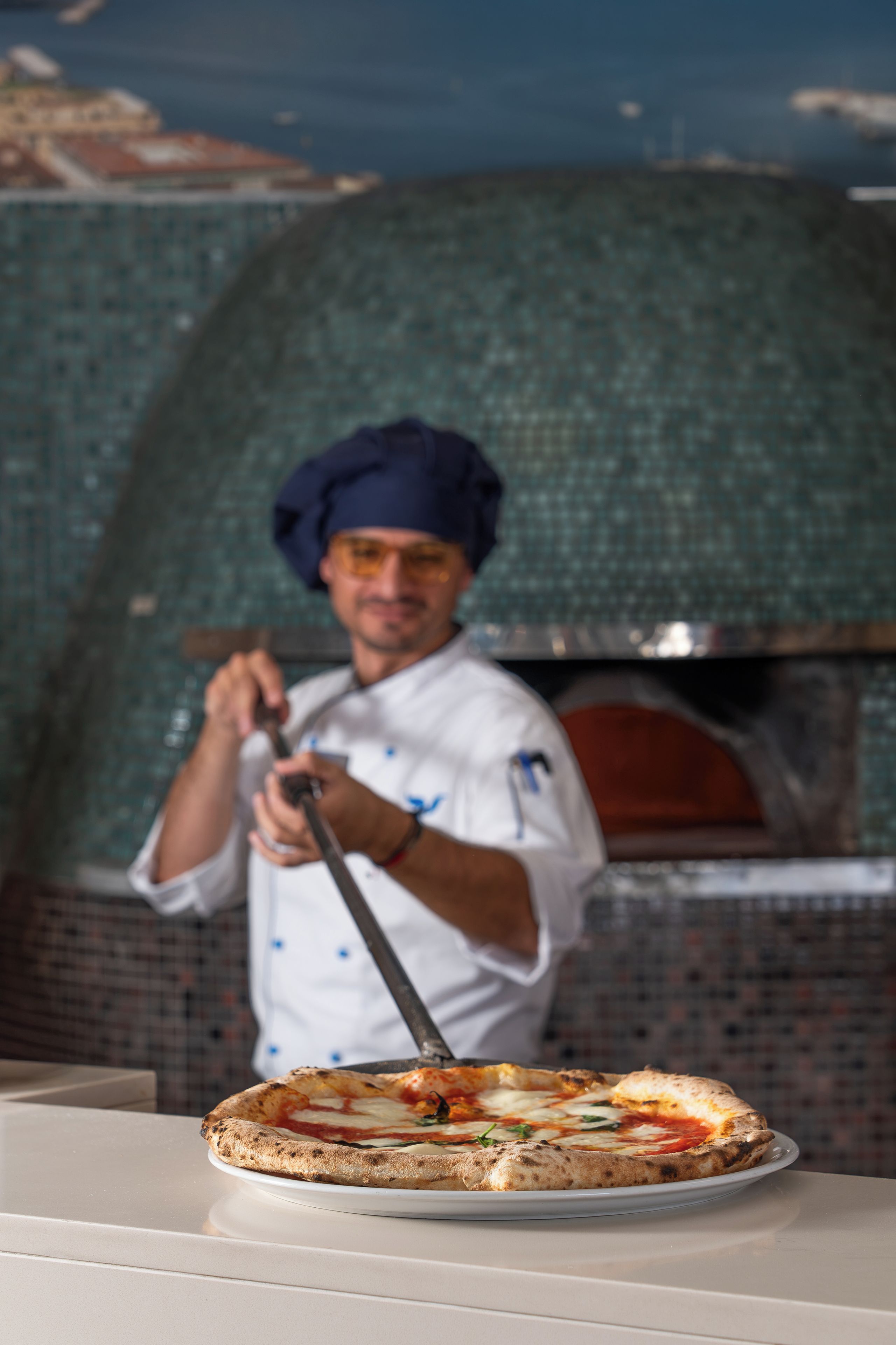 Un chef con uniforme blanco y gorro de cocinero, sosteniendo una pala para pizza frente a un horno de ladrillo.