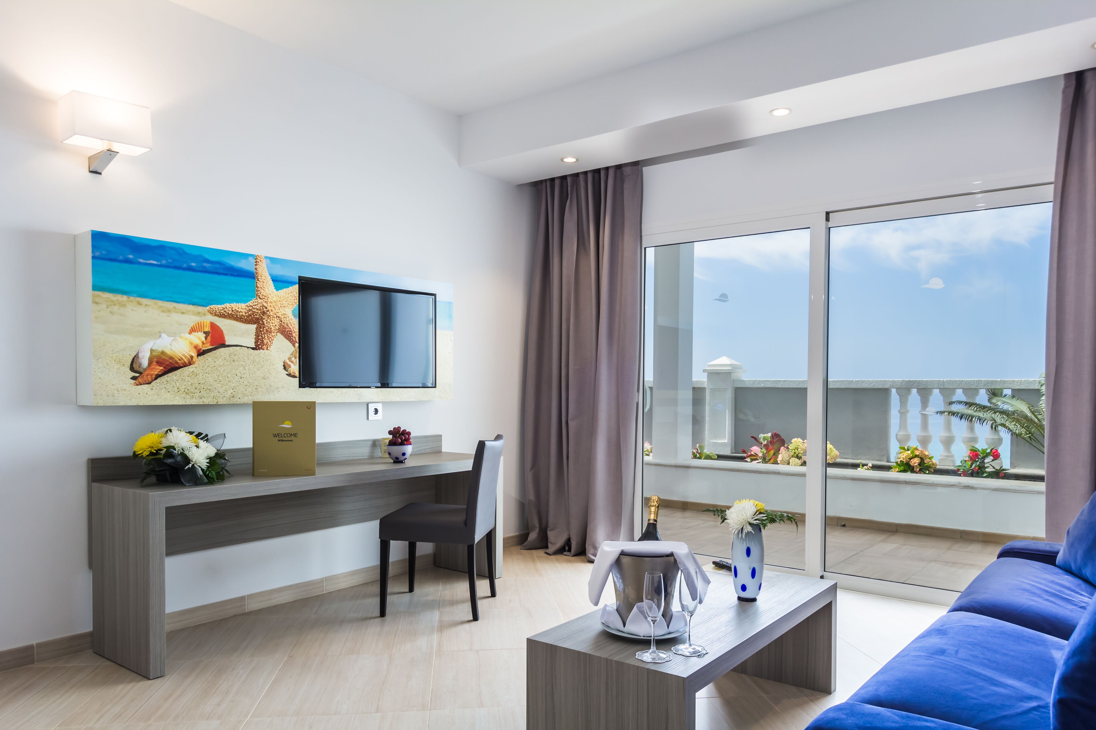 A modern living room with a flat-screen TV, a desk, a blue sofa, and a balcony with a view of the sea.
