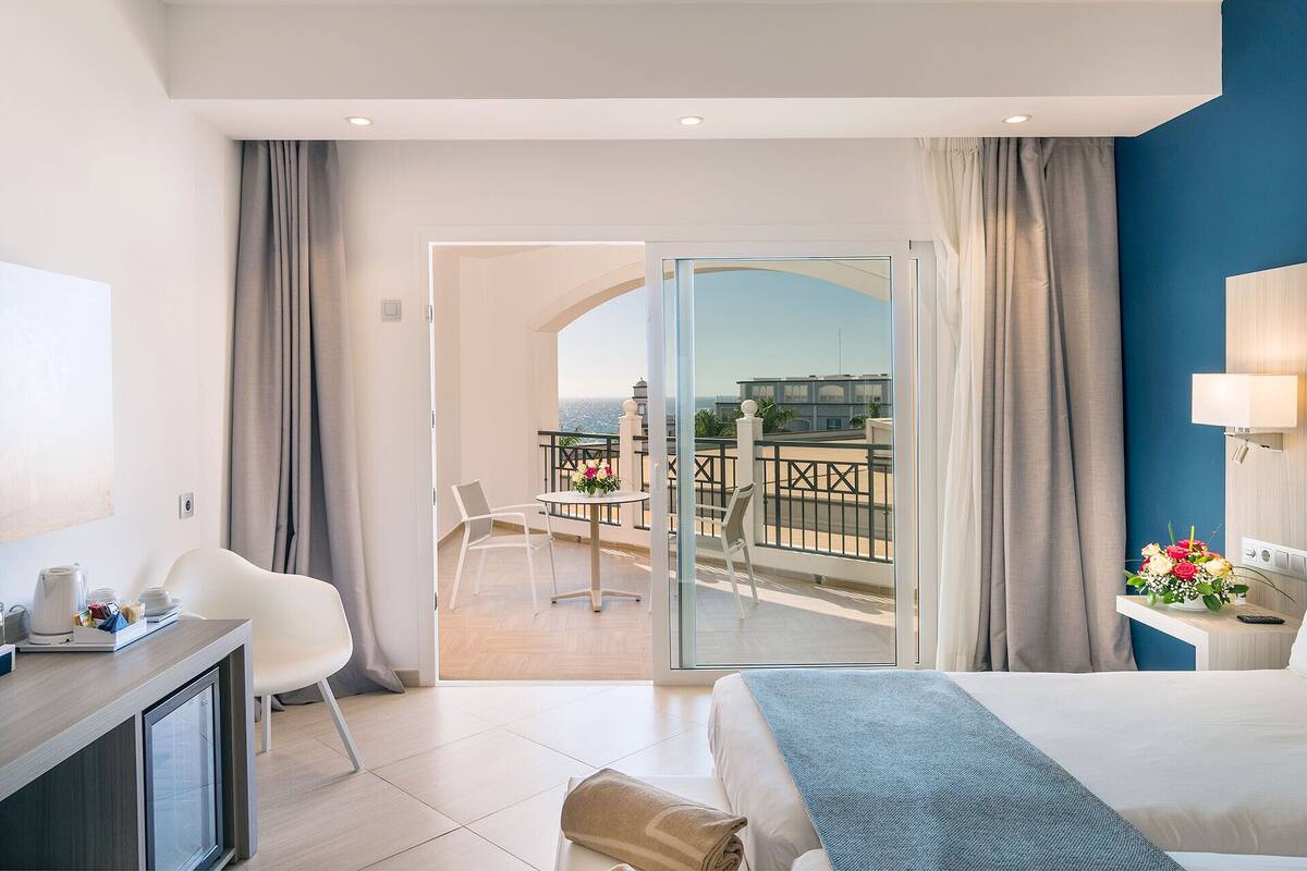 A hotel room with a bed, a desk, a chair, a balcony with a view of the city, and a window with curtains.