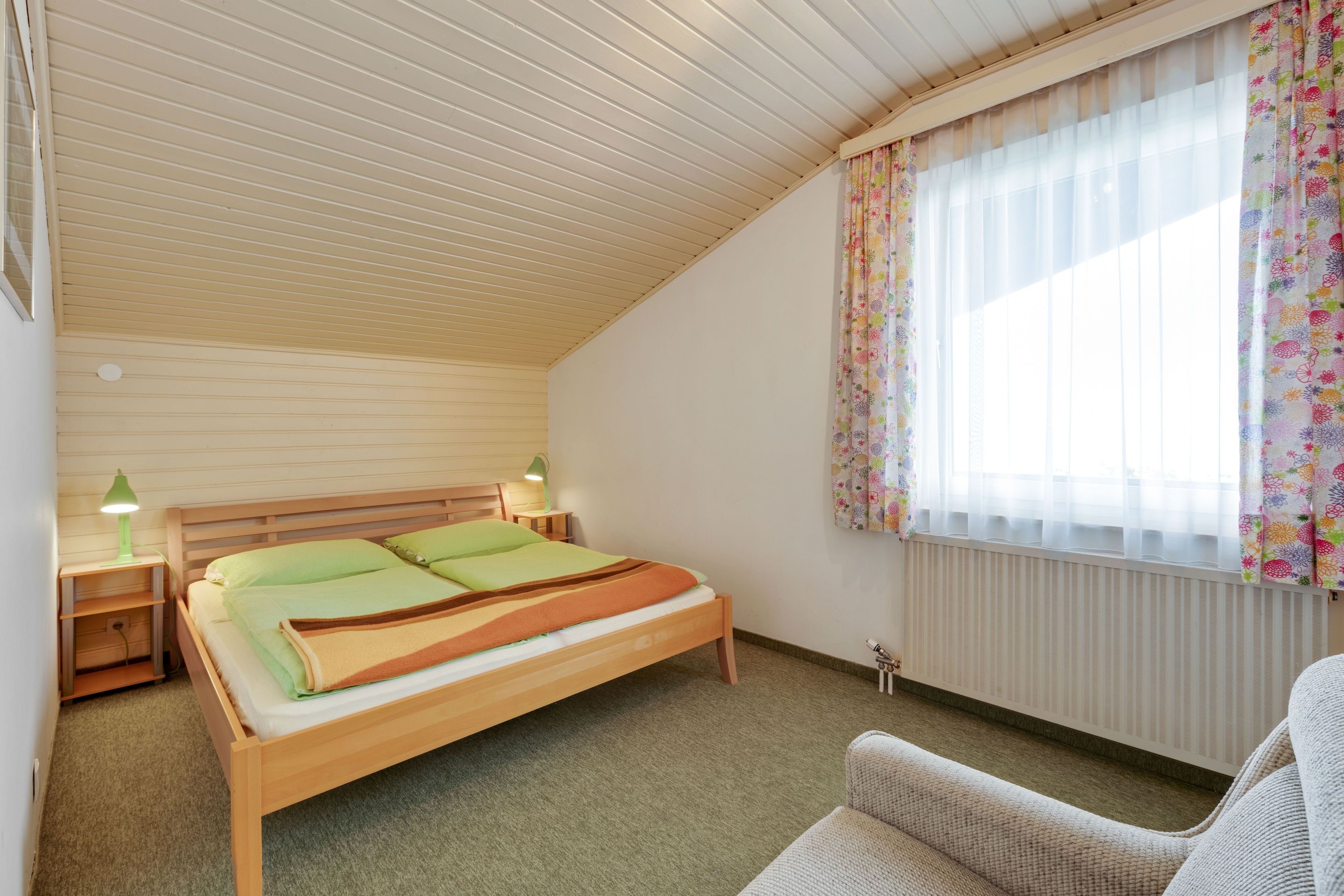 Un lit simple avec un cadre en bois, une table de chevet avec une lampe, un radiateur et une fenêtre ornée de rideaux à motifs floraux.