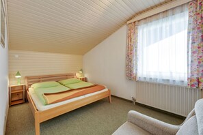 Una cama individual con estructura de madera, una mesita de noche con lámpara, un radiador y una ventana con cortinas florales.