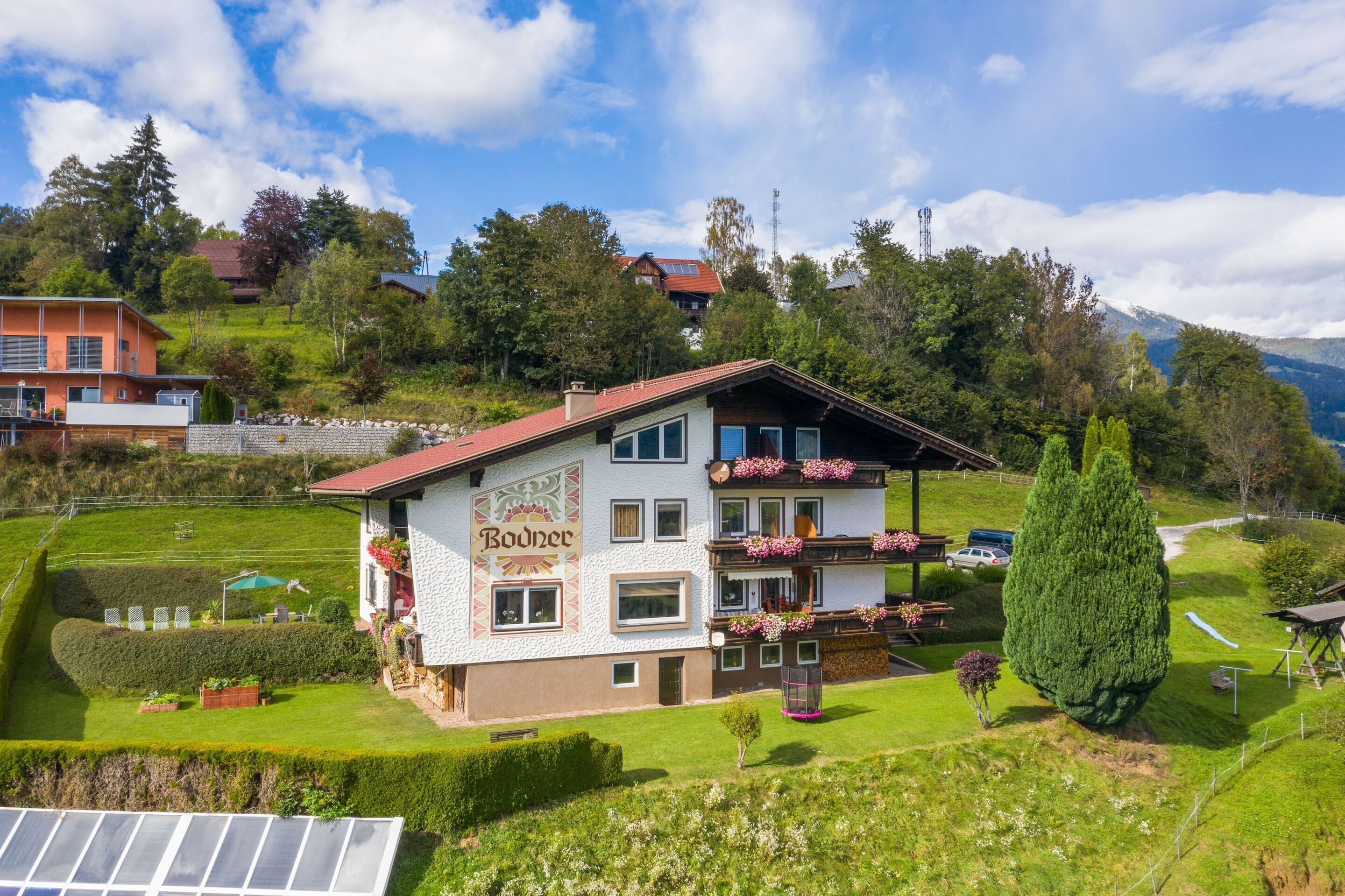A two-story building with a sign reading "Bodner" on the side, surrounded by a well-maintained garden and a view of a mountainous landscape.
