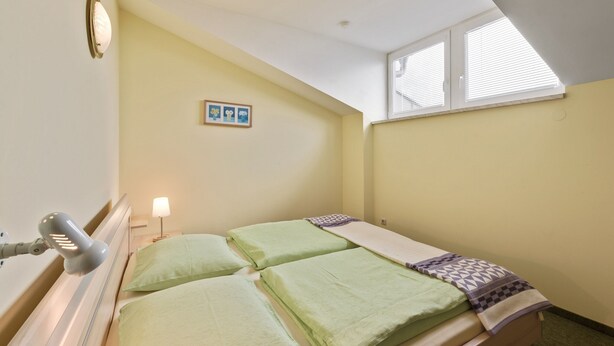 Ein Einzelbett mit grüner Bettwäsche, ein Nachttisch mit Lampe, ein Bild an der Wand und ein Oberlicht.