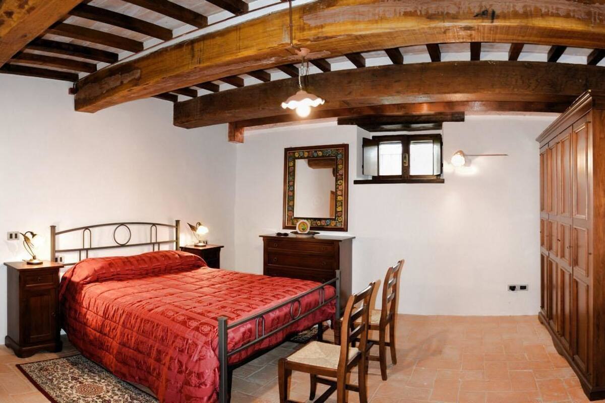 A bedroom with a wooden ceiling, a bed with a red bedspread, a wooden wardrobe, a bedside table with a lamp, and a mirror on the wall.