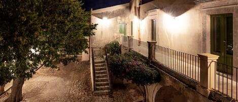 Une cour avec un chemin pavé, un arbre, un escalier et un bâtiment doté de portes vertes et d’un balcon.