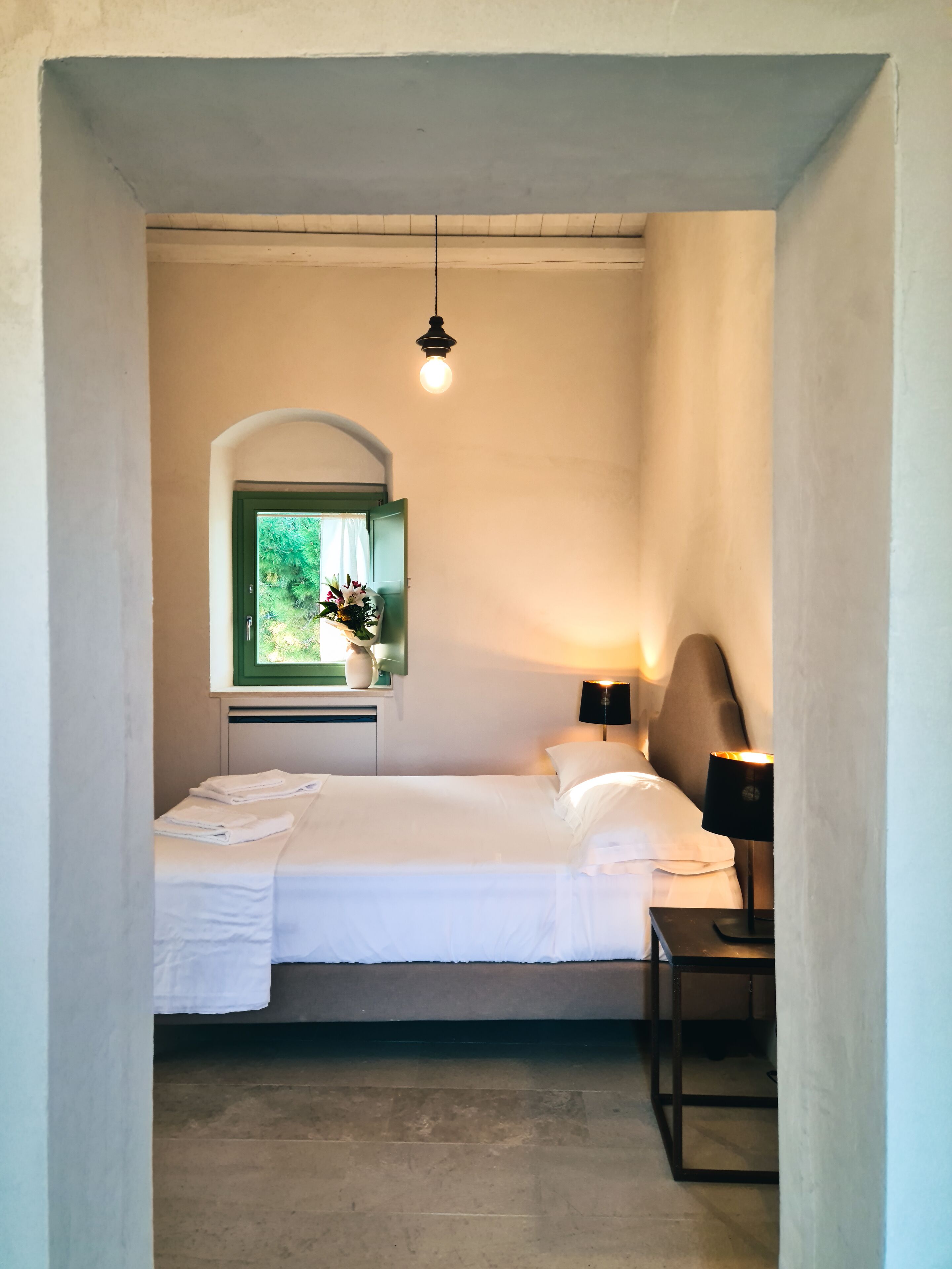 A bedroom with a bed, a bedside table, a lamp, a window with a view of greenery, and a hanging light fixture.