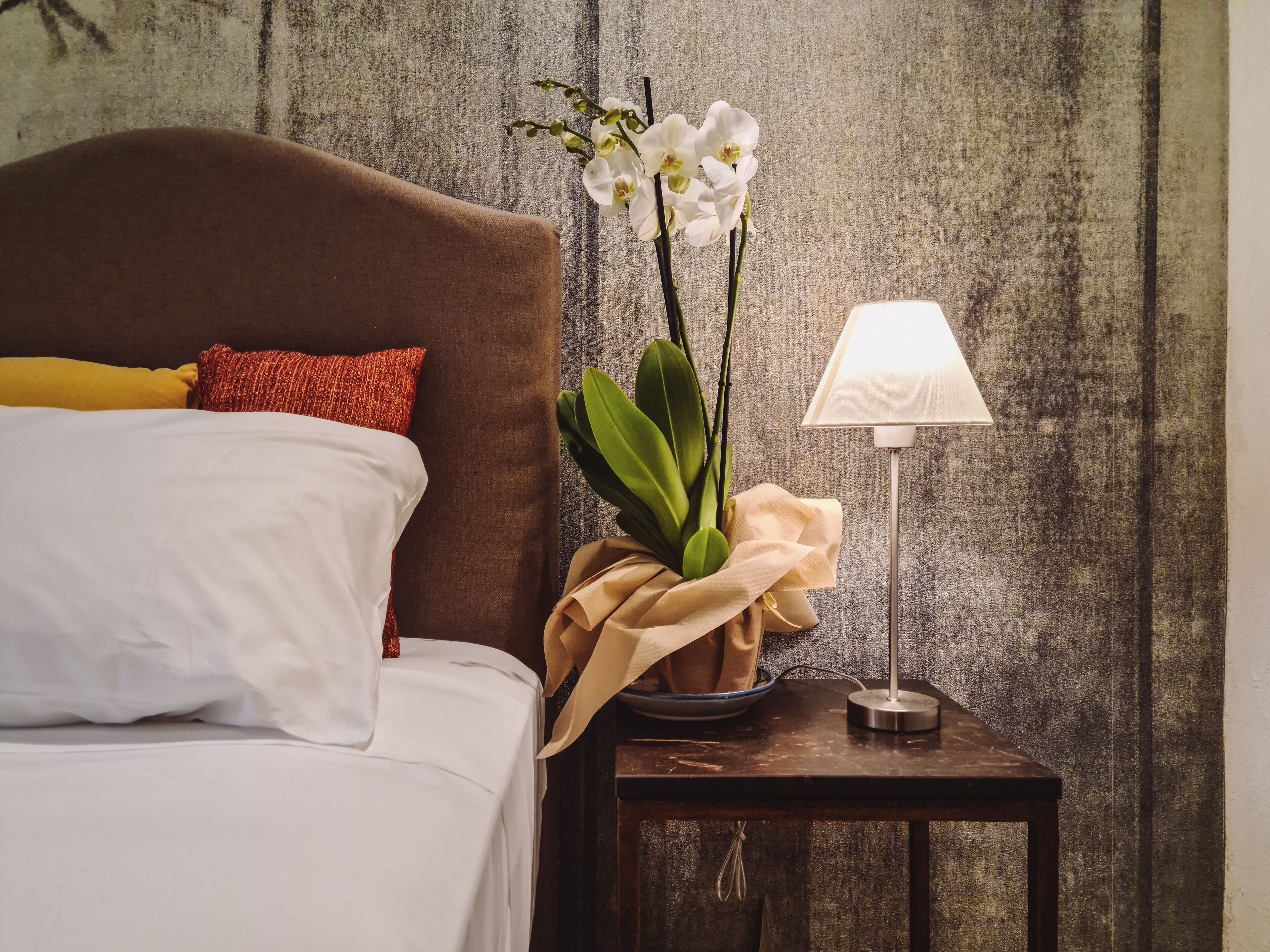 A neatly made bed with white and yellow pillows, a brown headboard, and a bedside table with a lamp and a bouquet of white orchids.