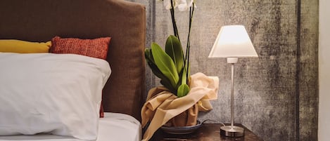 Cama bem arrumada com almofadas brancas e amarelas, cabeceira marrom e mesa de cabeceira com luminária e um buquê de orquídeas brancas.