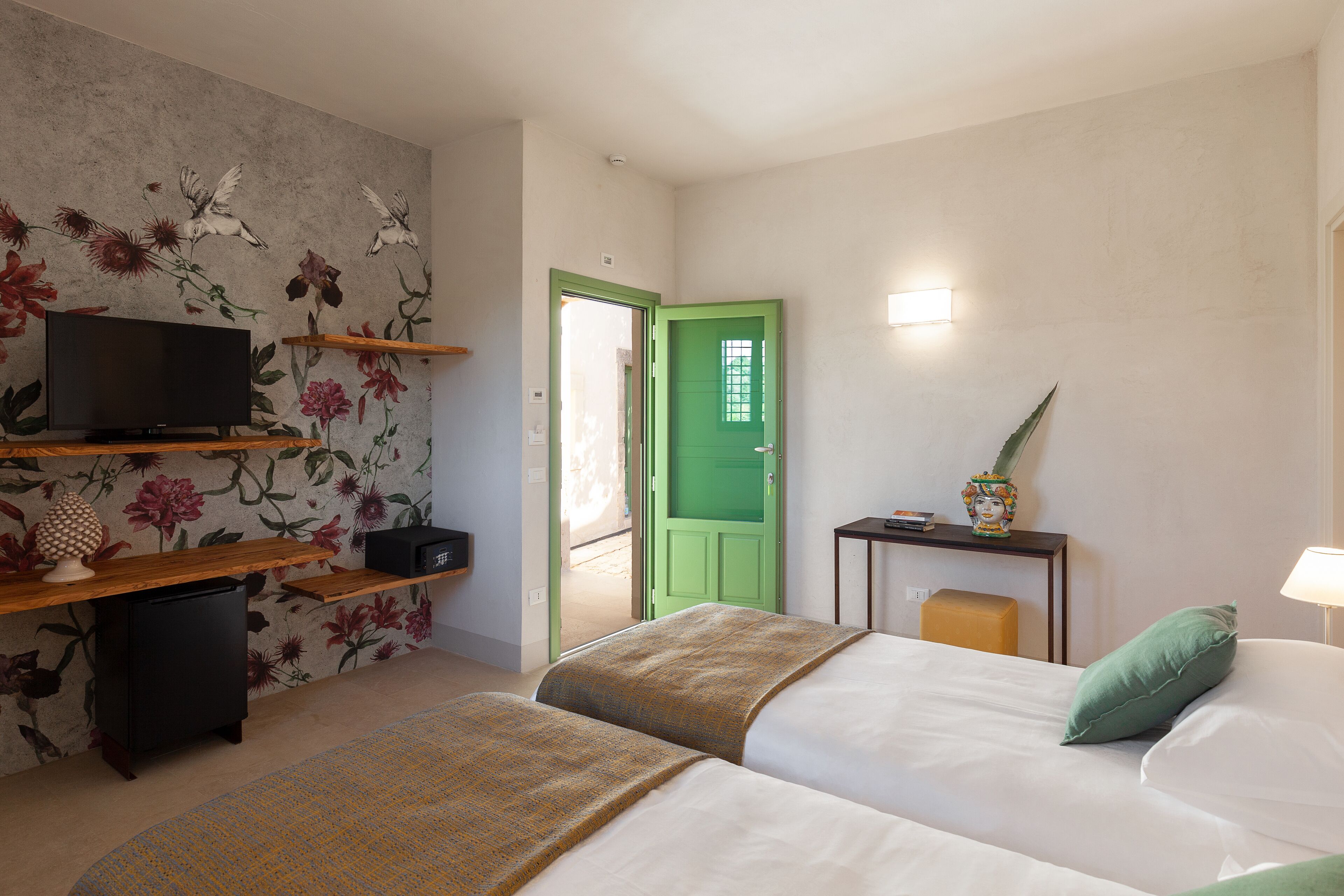 A hotel room with a bed, a TV on a shelf, a desk with a lamp, and a green door.
