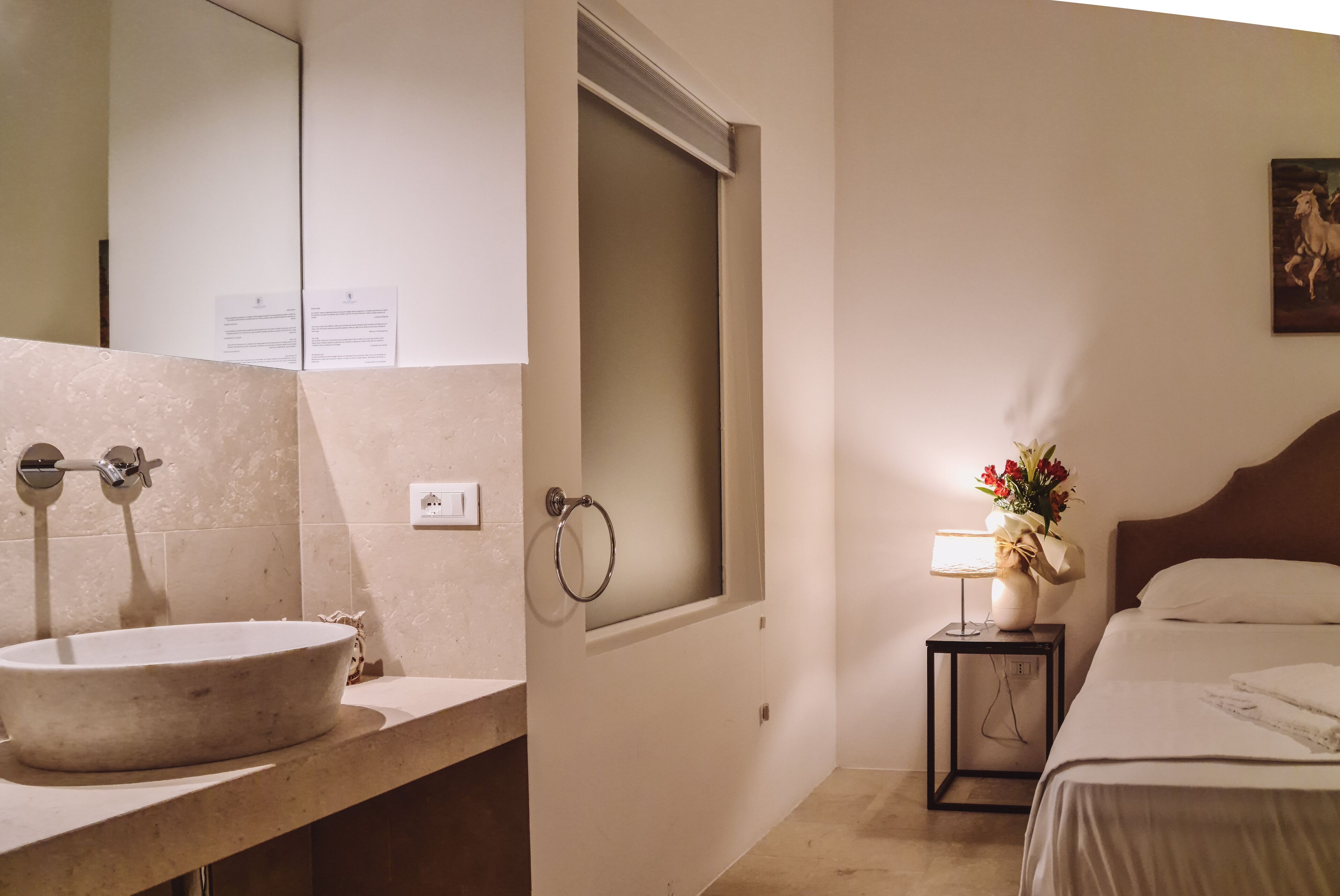 Un baño con un lavamanos grande, un espejo, un tozaleta y una mesita de noche con una lámpara y flores.
