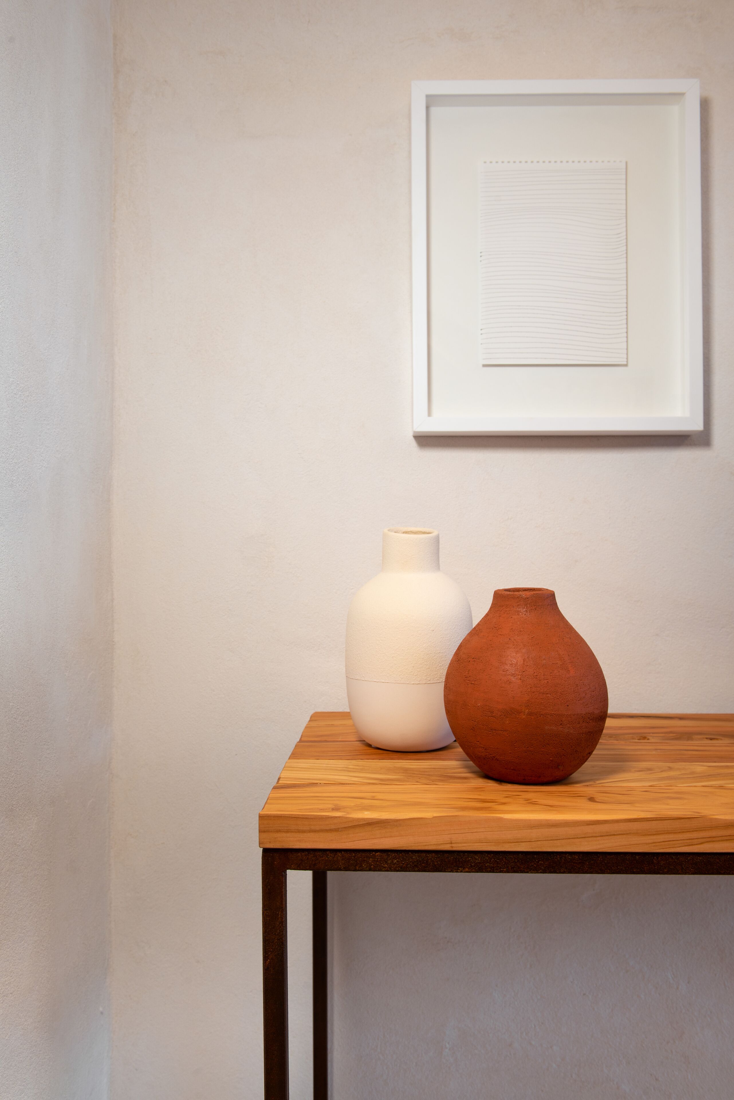 Ein Holztisch mit zwei Vasen, eine weiß und eine terrakottafarben, vor einer schlichten Wand mit einem gerahmten Bild darüber.