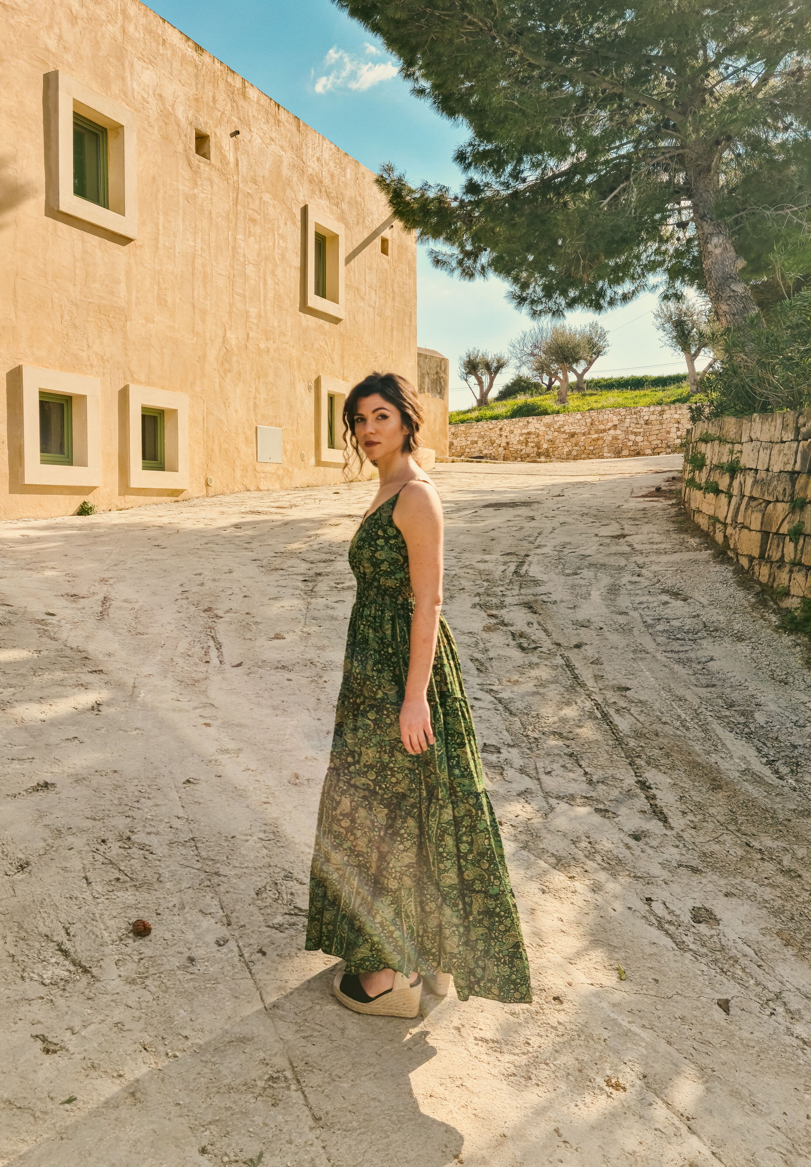 A woman in a patterned dress standing on a dirt path with a building in the background.