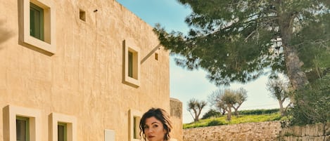 A woman in a patterned dress standing on a dirt path with a building in the background.