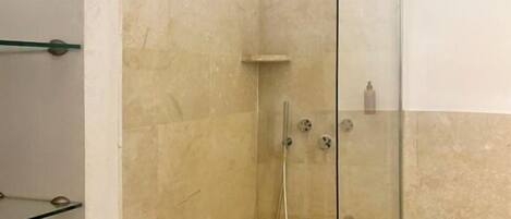 A glass-enclosed shower with a handheld showerhead, a wall-mounted faucet, and a small shelf with toiletries.