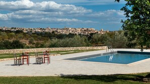 A swimming pool with a stone wall and chairs, a clear sky, and a distant cityscape. - Terre di Romanello Relais in Val di Noto (Noto)