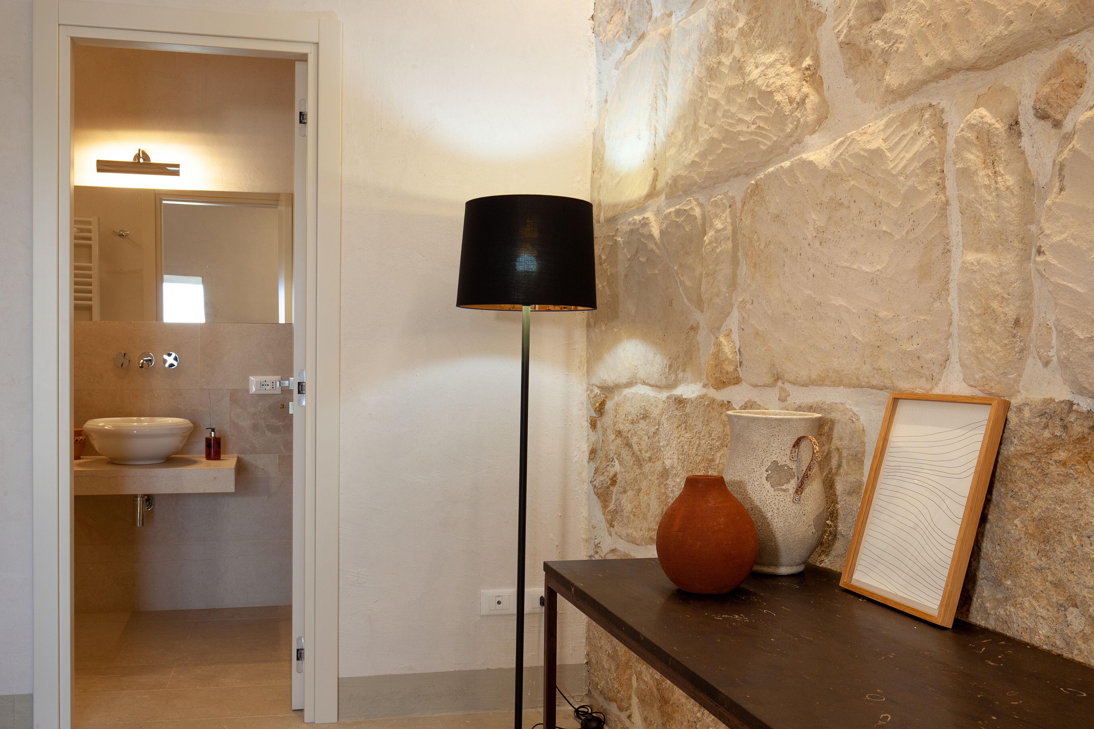 Un baño con un lavabo blanco, un espejo y una puerta que da a otra habitación. Hay una lámpara de pie negra y un florero en un estante.