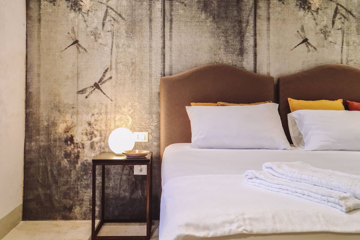 Una habitación de hotel con cama, mesita de noche con lámpara y pared texturizada con un mural de pájaros.
