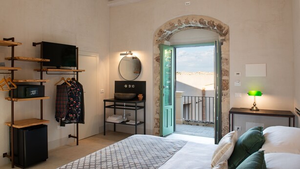 A hotel room with a bed, a desk with a lamp, a TV, a mirror, and a view of the outdoors. - Terre di Romanello Relais in Val di Noto (Noto)