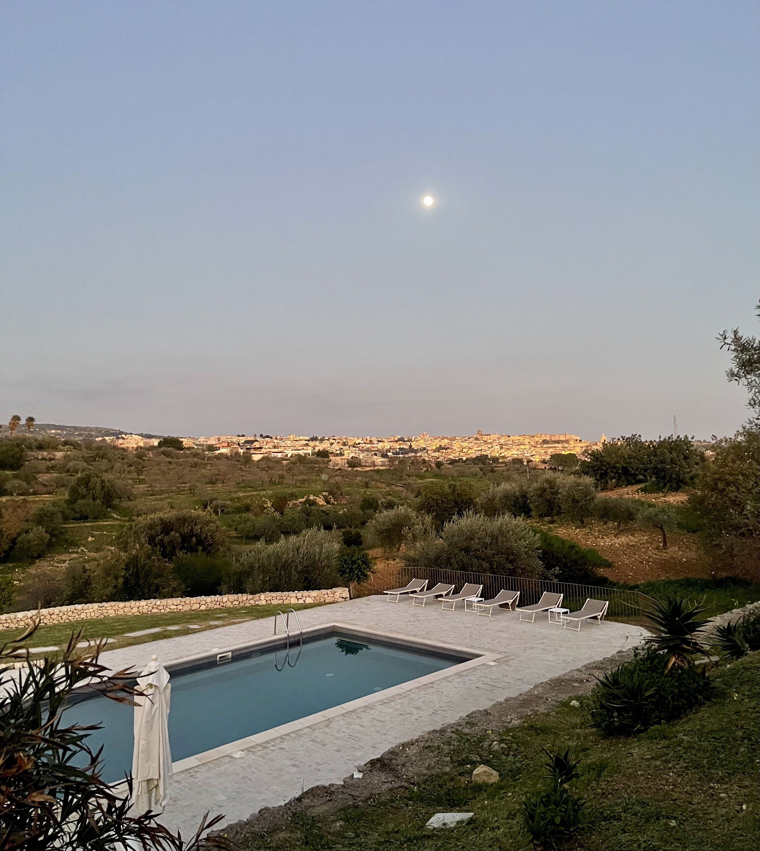 Una piscina con sillas de descanso, rodeada de follaje y un muro de piedra, con vistas a un pueblo lejano y un cielo despejado.