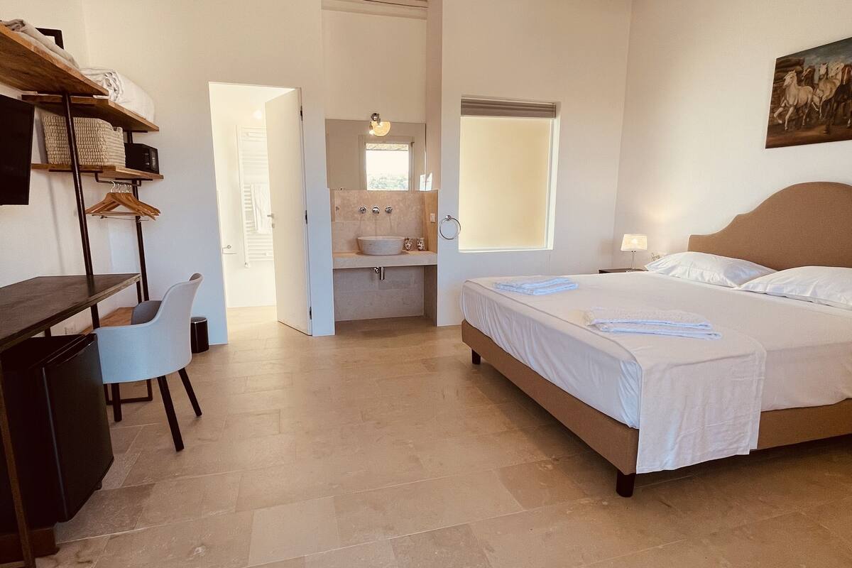 Un dormitorio con cama, escritorio y silla, baño con lavabo y espejo, y ventana.