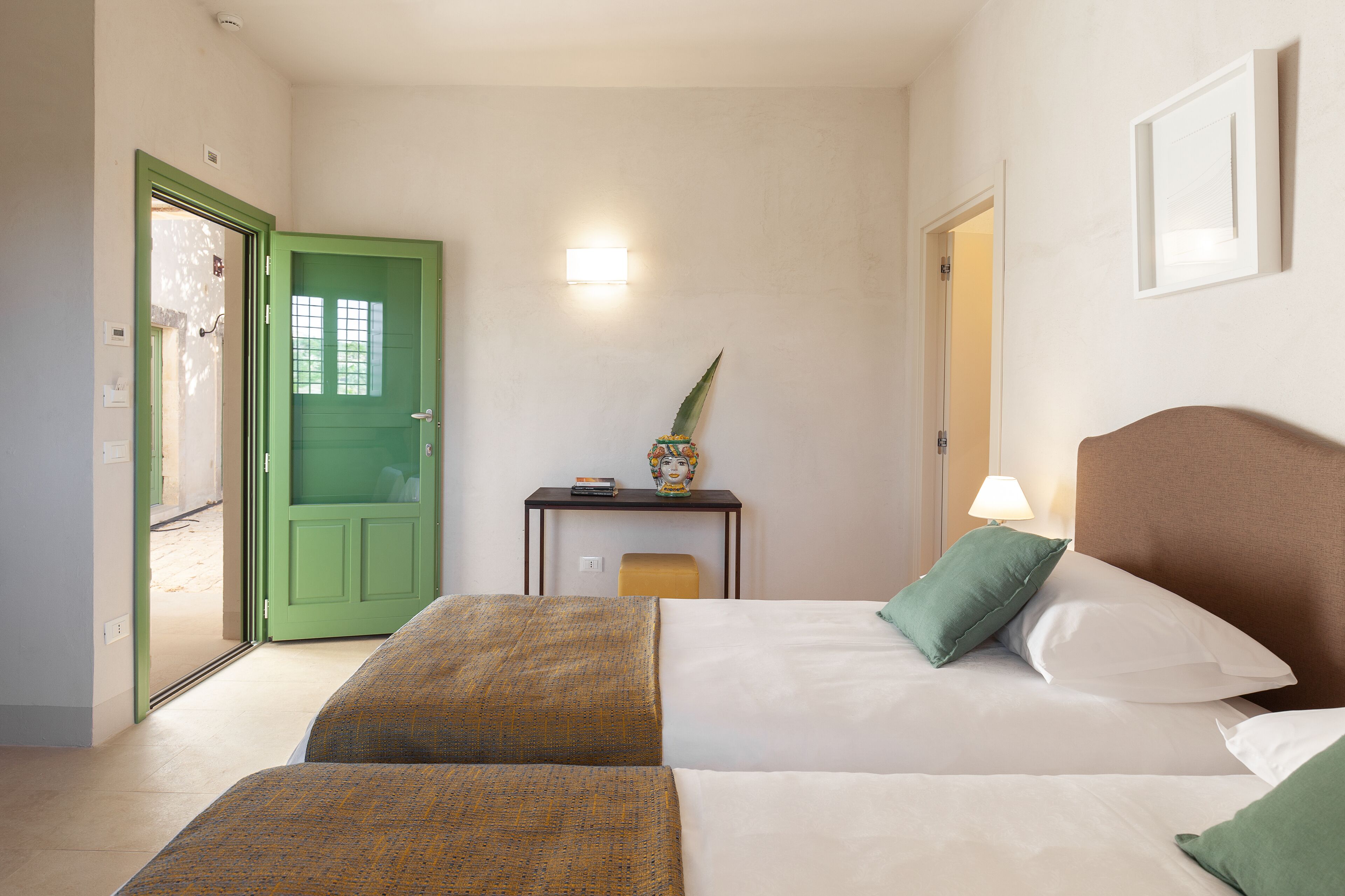 Quarto de hotel com duas camas, uma porta verde, uma mesinha com luminária e uma luz na parede.
