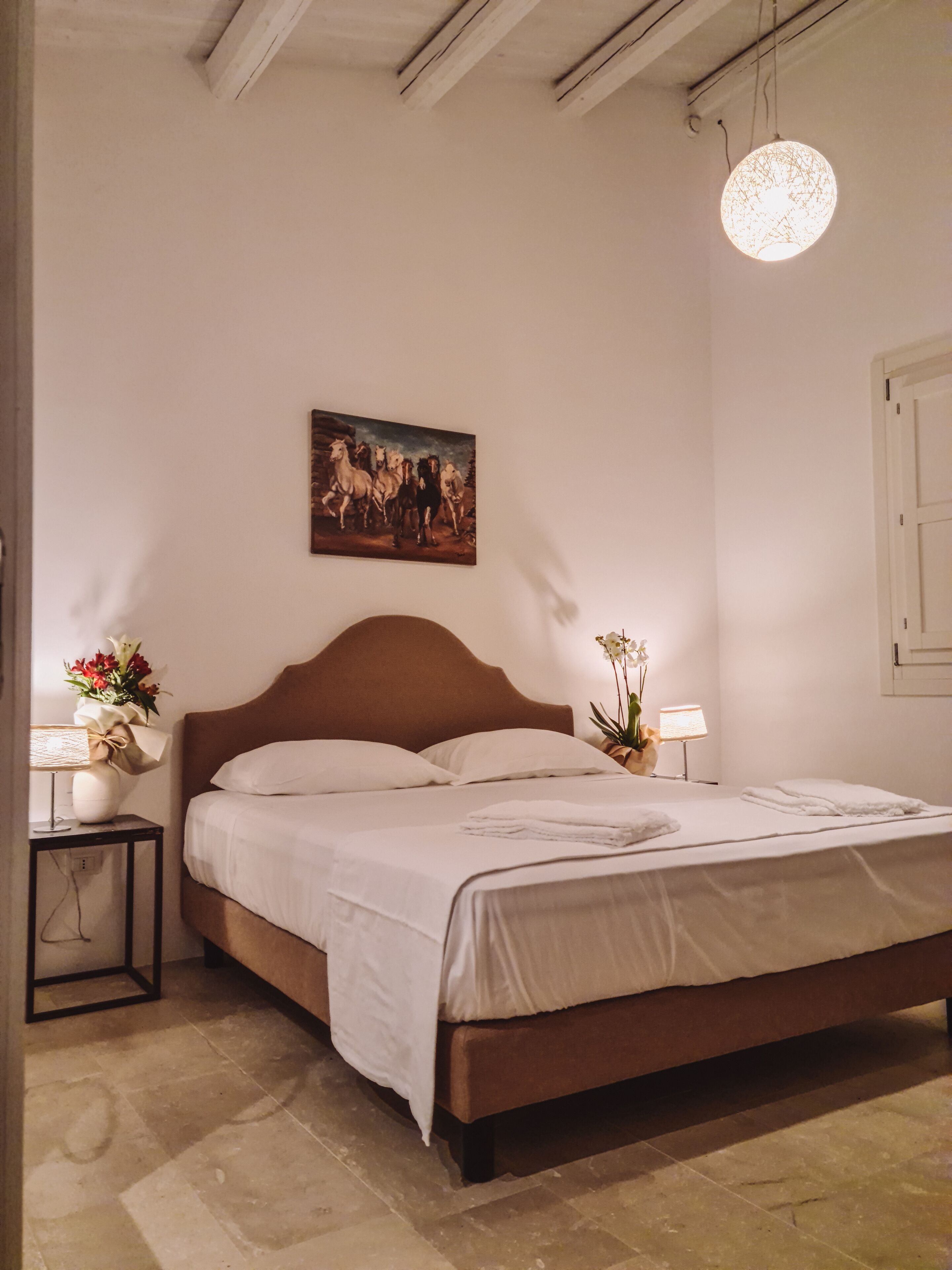 Une chambre à coucher avec une tête de lit en bois, un tableau représentant des chevaux accroché au mur, une table de chevet avec une lampe et une plante en pot.